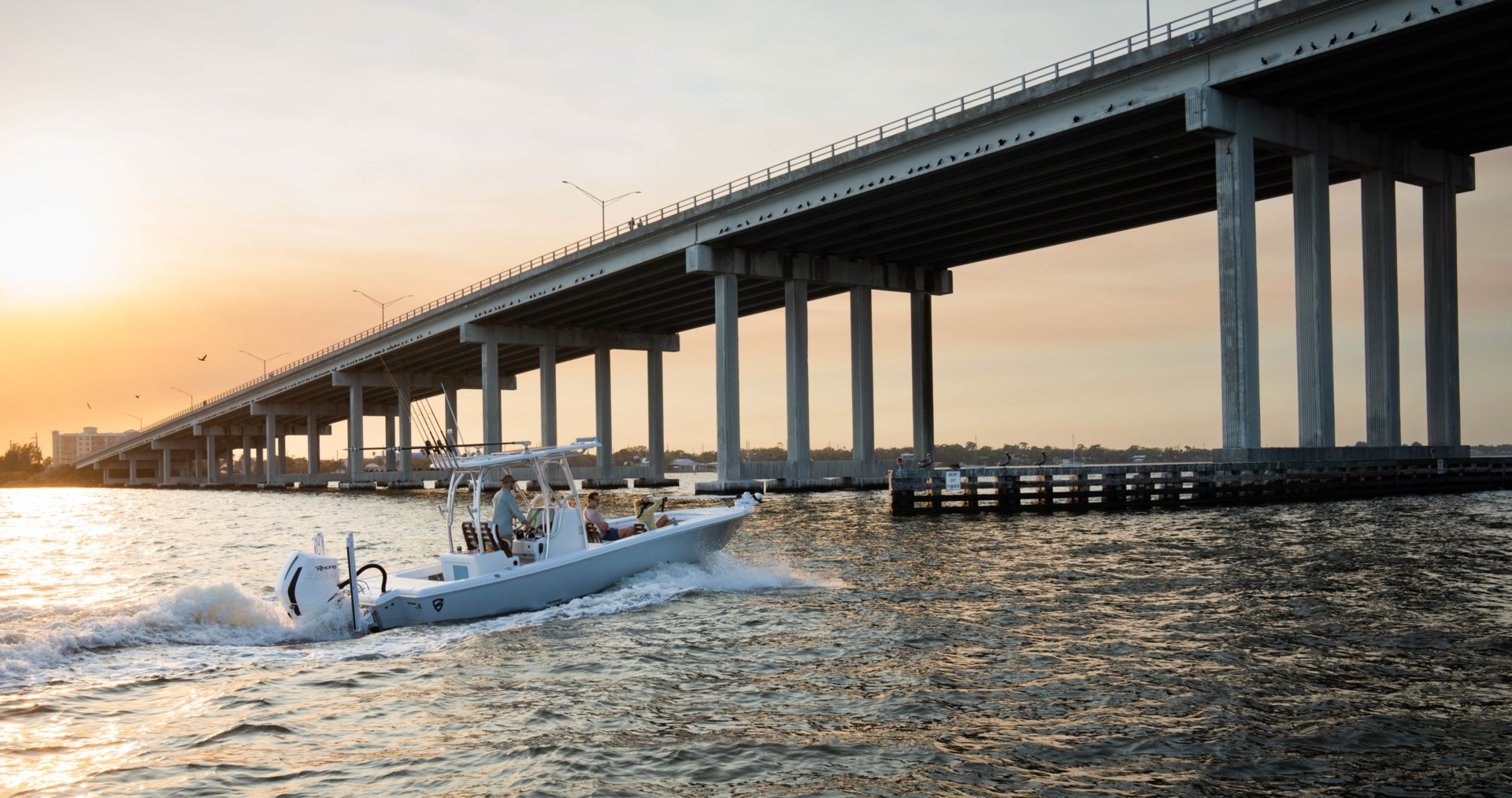 A white center console fishing boat passing under a bridge with a setting sun in the background