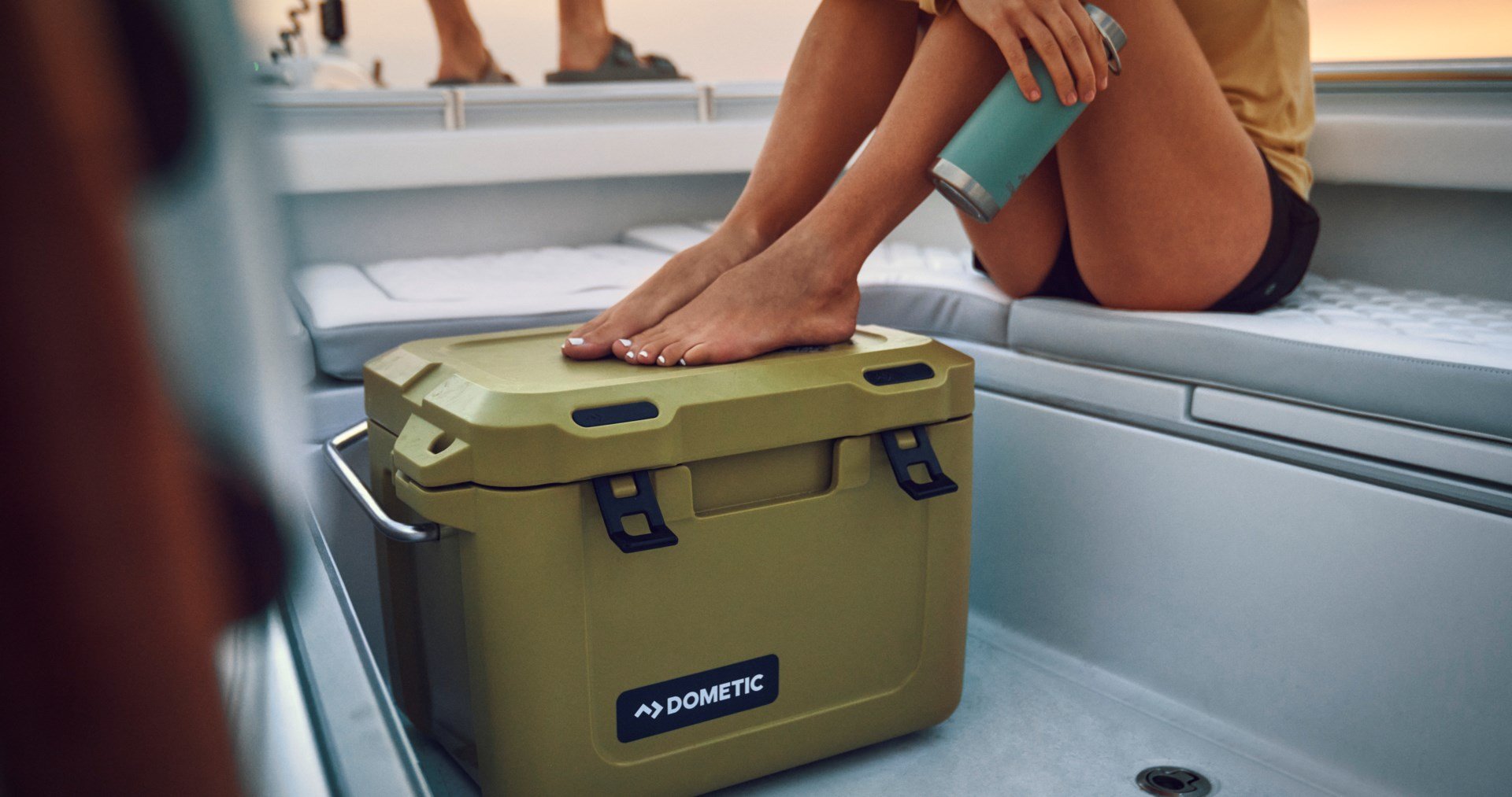 An olive colored Dometic Patrol  20 cooler on a boat next to a woman drinking from a Dometic drinkware tumbler